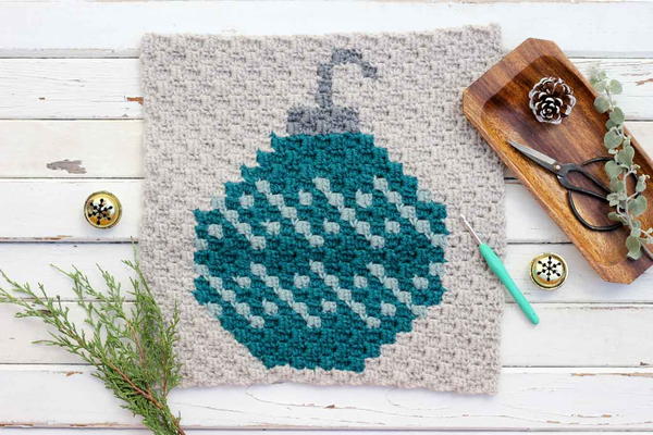 Image shows the C2C Christmas Bulb Afghan/Pillow Square on a rustic wood background and accessories surrounding it.