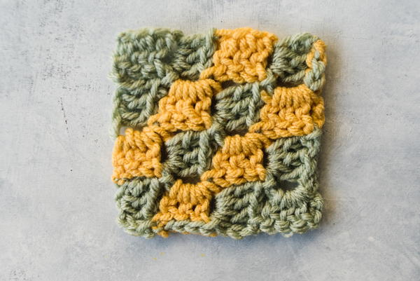 What is C2C Crochet? Image shows a corner to corner swatch in green and yellow yarn and on a marbled light gray background.