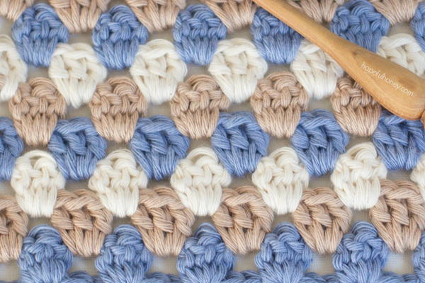 Image shows a close up of a granny stripe crochet swatch in muted brown, blue, and white. There is a wooden crochet hook at the top right.