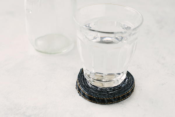 Image shows the finished rolled jean coaster sitting on a table with a glass on top. There is also a glass behind without a coaster.
