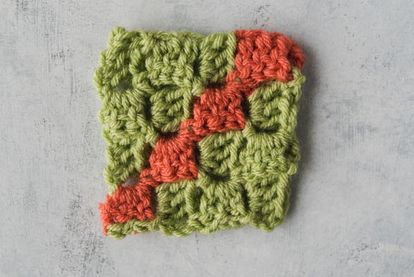 Image shows a corner to corner swatch in green and coral yarn and on a marbled light gray background.