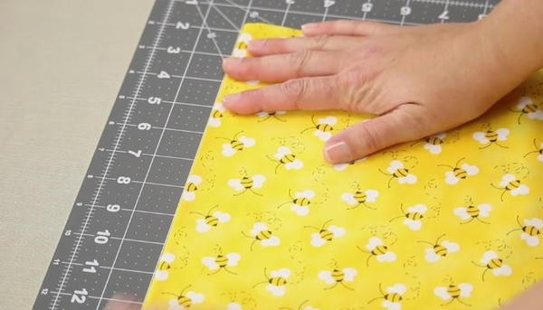 Image shows a close up of a gray cutting mat on a beige table. A hand is adjusting a yellow fabric with bees on the mat.