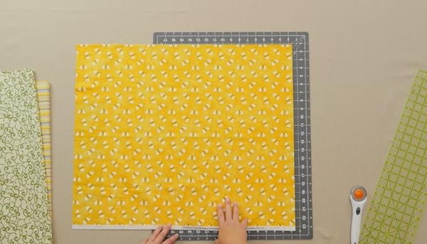 Image shows an overhead view of a gray cutting mat on a beige table. There is a stack of fabric on the left and a ruler and rotary cutter on the right. Hands are adjusting a yellow fabric with bees on the mat.