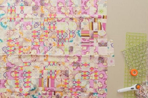 Image shows several quilt blocks sewn together on a cream background. There's a couple rulers, glow tape, and a rotary cutter off to the right side.