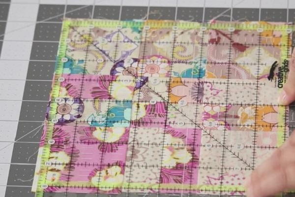 Image shows a quilt block on a cutting mat with a quilting ruler laid on top. Hands are adjusting the block and ruler to match up as needed.