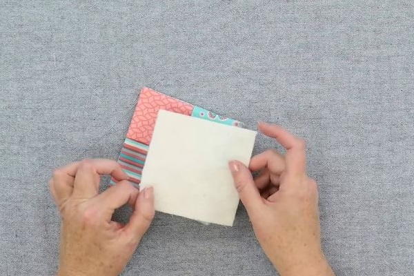 Image shows hands holding a square of cotton batting above the sewn folded fabric coaster on a gray background.