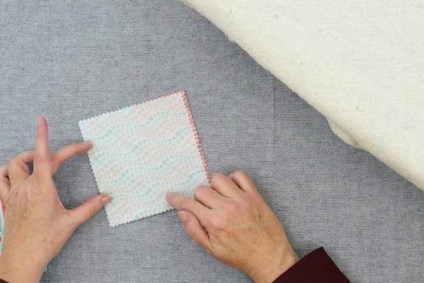 Image shows hands putting the backing square of fabric on the back of the square folded fabric coaster on a gray background.