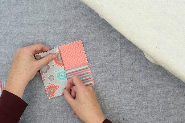 Image shows hands arranging four rectangle fabric pieces to make the square folded fabric coaster on a gray background.
