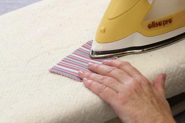 Image shows an iron pressing down a rectangle piece of fabric on an ironing board.