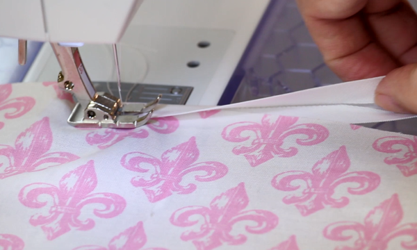 Image shows a close-up of a sewing machine sewing bias tape over the edges of white fabric with pink fleur-de-lis designs.