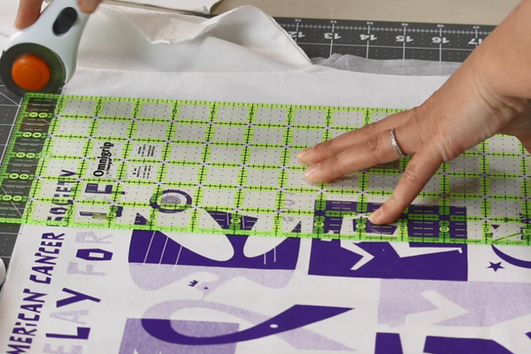 Image shows a shirt quilt block up close with a ruler over it and a rotary cutter starting to cut.