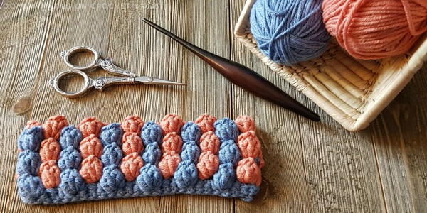 Image shows the finished popcorn stitch swatch in blue and pinkish orange. There is a crochet hook, pair of scissors, and a basket with two balls of yarn.