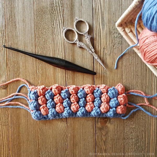 Image shows the nearly popcorn stitch swatch in blue and pinkish orange. The yarn ends are still loose. There is a crochet hook, pair of scissors, and a basket with two balls of yarn.
