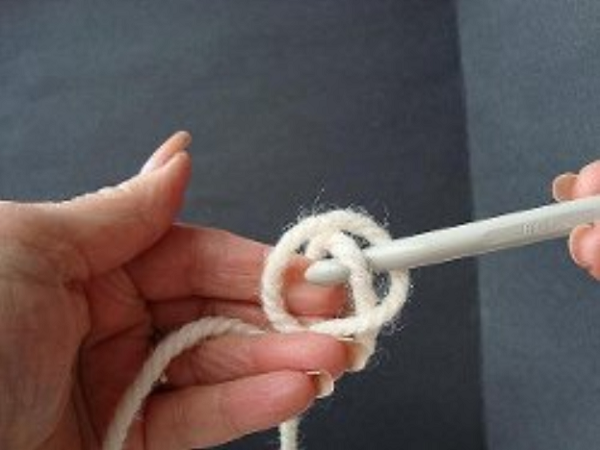 Image shows step 2 for crocheting a magic ring, which shows a hand holding yarn in a loop with a hook in the other hand pulling the yarn through.