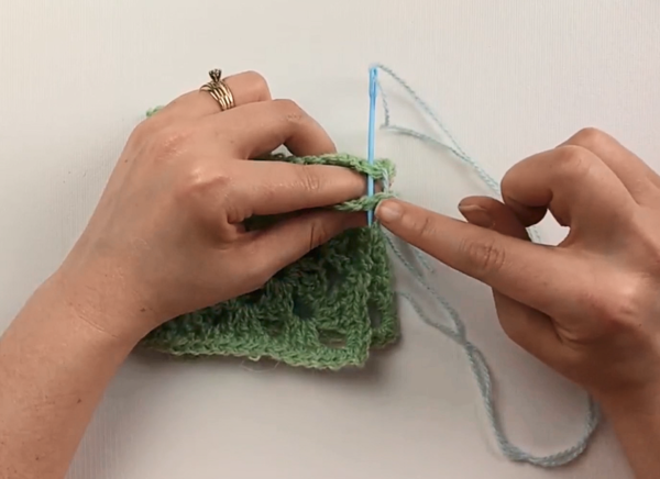Image shows two hands whip stitching two green granny squares together with a blue yarn needle and yarn.