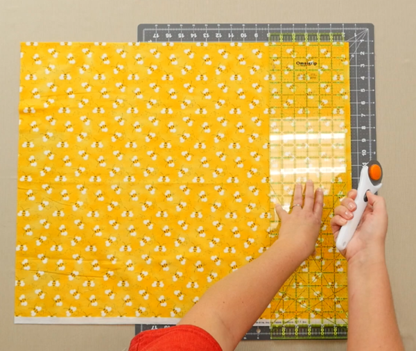 Image shows an overhead view of a gray cutting mat on a beige table. One hand is adjusting a clear quilting ruler over the yellow fabric with bees on the mat. The other hand is holding a rotary cutter.