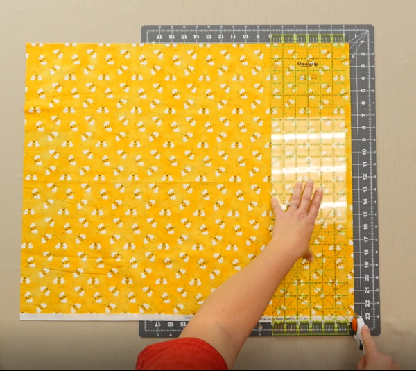 Image shows an overhead view of a gray cutting mat on a beige table. One hand is holding down a clear quilting ruler over the yellow fabric with bees on the mat. The other hand is holding the rotary cutter down at the bottom edge of the fabric and ruler, 