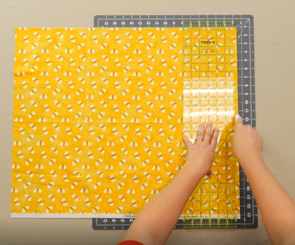 Image shows an overhead view of a gray cutting mat on a beige table. One hand is holding down a clear quilting ruler over the yellow fabric with bees on the mat. The other hand is holding the rotary cutter, cutting up about halfway to the top of the fabri