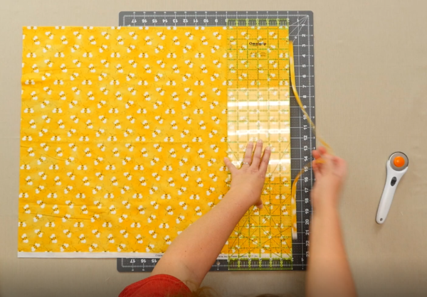 Image shows an overhead view of a gray cutting mat on a beige table. One hand is holding down a clear quilting ruler over the yellow fabric with bees on the mat. The other hand is pulling off the cut excess fabric. The rotary cutter is sitting off to the 