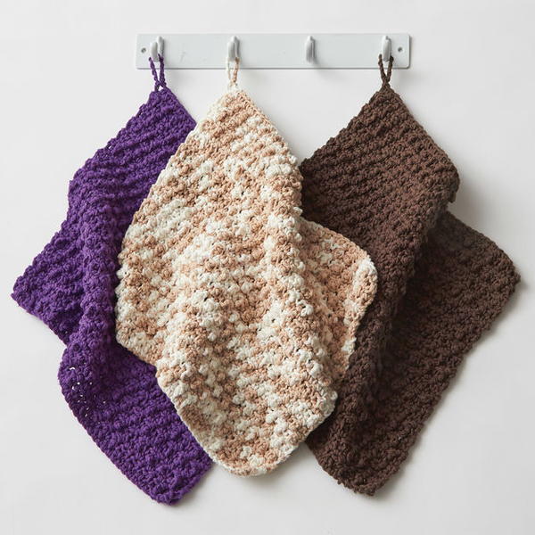 Image shows three dishcloths hanging on a bar in purple, beige variegation, and brown. These are the Super Speedy Textured Dishcloth pattern.