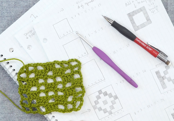 Image shows a gray carpet background with notebook and piece of loose paper with filet crochet designs and notes. On top of the paper is a small green filet crochet swatch, a crochet hook, and mechanical pencil.