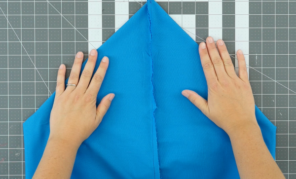 Image shows a cutting mat in the background. The sewn blue bag piece is being pushed flat by two hands.