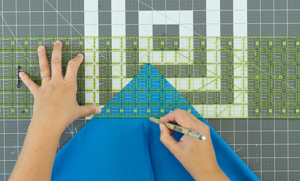 Image shows a cutting mat in the background. The sewn blue bag piece is being measured by a quilting ruler and a person is marking the measurement needed with a fabric marker.