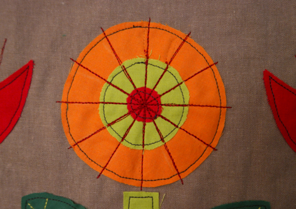 Image shows close-up of an orange embroidered flower with 12 lines added through the center, on the cushion. How to Embroider the Flowers - Step 3