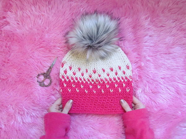Image shows the Faded Tunisian Beanie sitting on a pink fur background with hands holding the bottom. There are scissors set at the left of the hat.