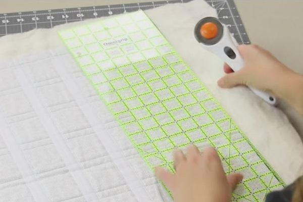 Image shows the piece of light fabric with all of the Velcro pieces attached sitting on a cutting mat. A ruler is sitting above and a hand is holding a rotary cutter in preparation of cutting the fabric.
