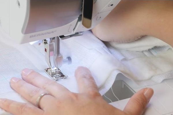 Image shows the Velcro being sewn with a machine to the piece of light fabric. A hand is holding the fabric still.