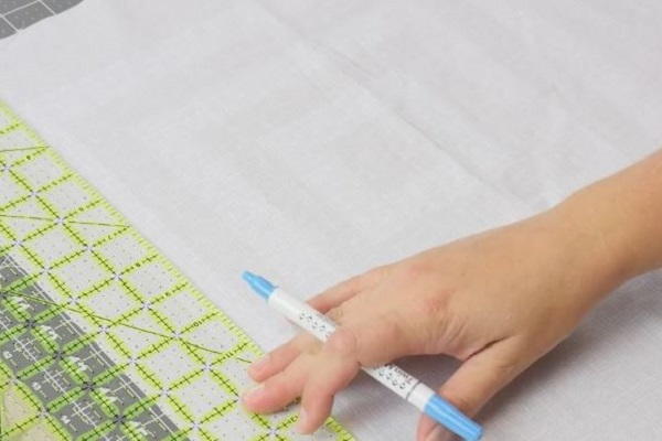 Image shows a piece of light fabric being measured with a quilt ruler. A hand is holding a fabric marking pen. Image shows a piece of light fabric being measured with a quilt ruler. A hand is holding a fabric marking pen.
