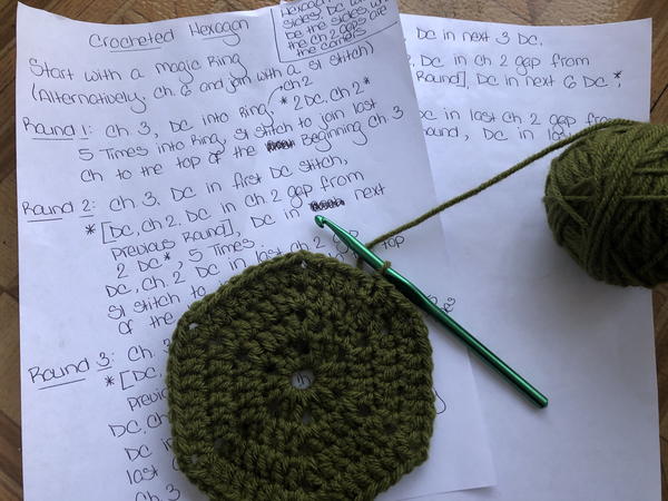 Image shows two pieces of printer paper with a crochet pattern written down. Lying on the paper is a dark green crochet hexagon in progress with a crochet hook and a ball of yarn attached.