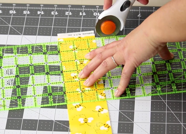 Image shows a close up of a gray cutting mat on a beige table. A hand is using the rotary cutter to cut the fabric at the edge of the ruler on the mat.