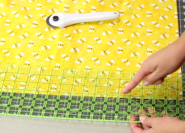 Image shows a close up of a gray cutting mat on a beige table. A hand is adjusting the quilting ruler over the yellow fabric on the mat. Another hand is pointing to a measurement on the ruler. The rotary cutter is sitting on the fabric.