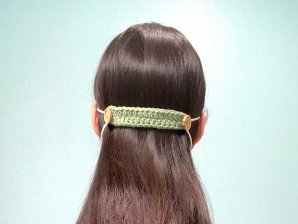 Image shows the back of a woman's head with a light green crochet ear saver holding elastic loops of a mask.