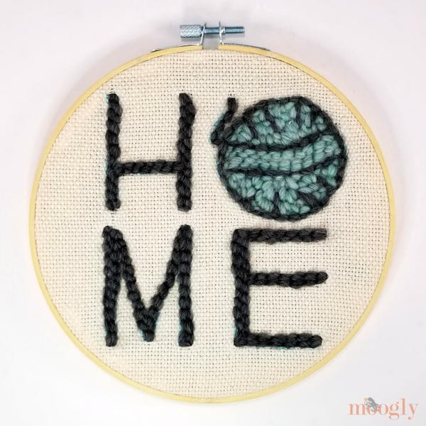 Image shows a punch needle embroidery design spelling out "HOME" with yarn as the "O". It is inside a unfinished wood embroidery hoop.