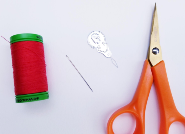 Image shows a spool of red thread, a needle, a needle threader, and scissors on a light background.