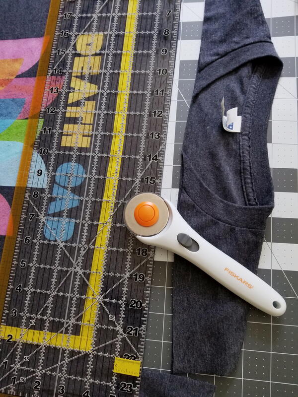 Image shows a shirt with the top cut and pulled away from the rest. It is on a mat with a ruler and rotary cutter laid on top of the shirt.