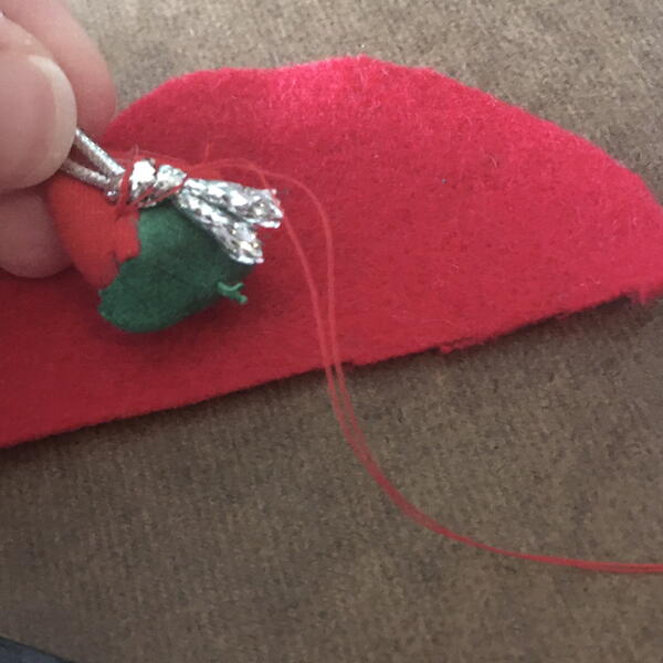Image shows the half-circle of red felt, and the pincushion strawberry being sewn to the silver string for the DIY Gnome Ornament.