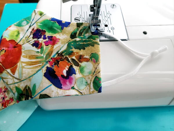 Image shows a sewing machine sewing the earloops onto the floral fabric face mask.