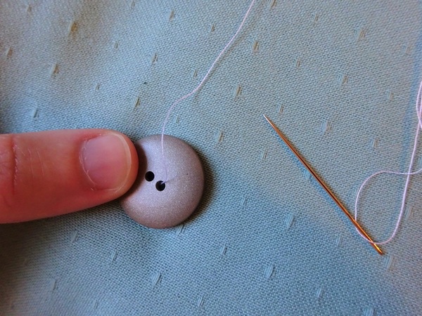 Image shows the hand sewing needle threaded and a button being held down by a thumb on fabric.