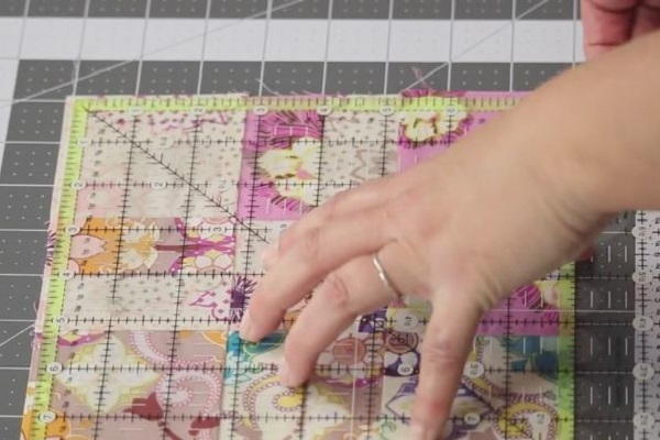 Image shows a quilt block on a cutting mat with a quilting ruler laid on top. One hand is holding down the ruler.