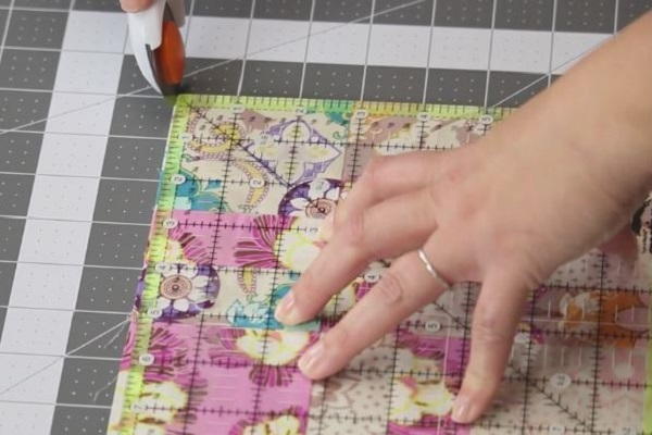 Image shows a quilt block on a cutting mat with a quilting ruler laid on top. One hand is holding down the ruler and the other is cutting the excess off with a rotary cutter.