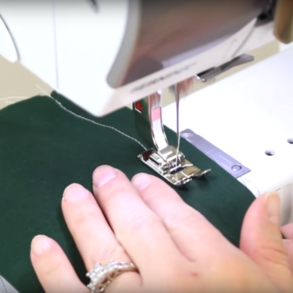 Image shows a sewing machine sewing a straight stitch on fabric, with a hand guiding the fabric.