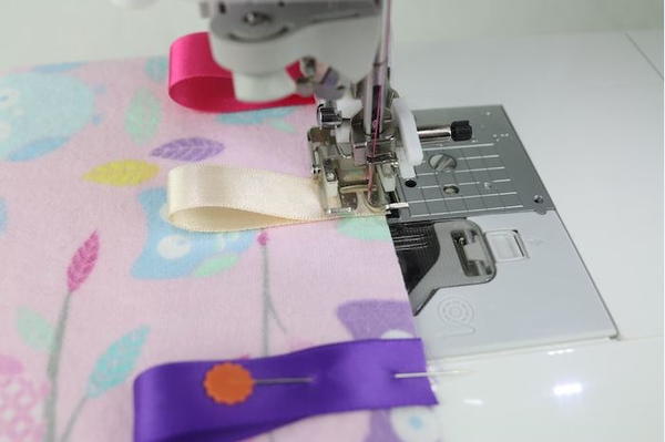 Image shows a sewing machine sewing the folded and pinned ribbons to the fabric piece.