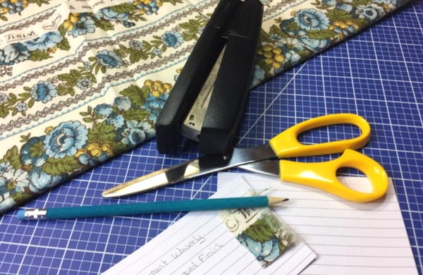 Fabric, stapler, scissors, pencil, and notecards on a cutting mat