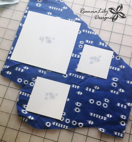 Planning Your Scrap Placement: Image shows a piece of fabric on a cutting mat. On top of the fabric is three different-sized paper squares for planning cuts.