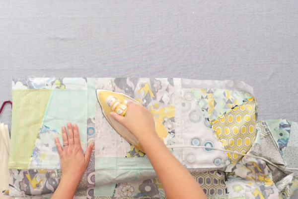 Image shows hands flattening and ironing the quilt top on an ironing board on gray surface.