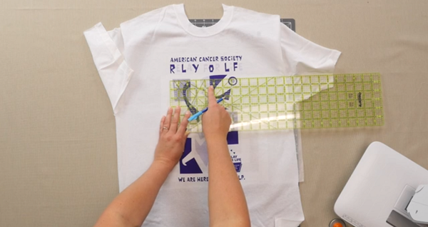 Image shows shirt on a table. Hands are using a ruler to measure the shirt design.